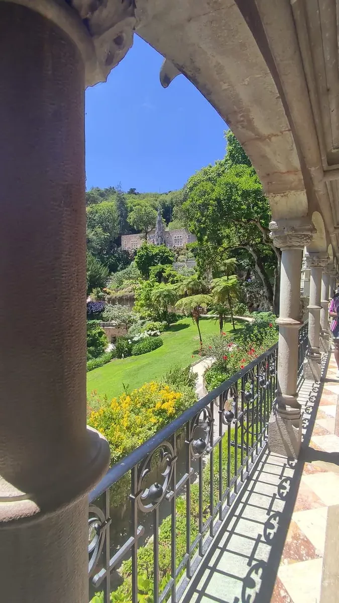 View from Quinta da Regaleira balcony over manicured gardens with palm trees on a sunny day. The columns lining the balcony are just one of the ornate decorations you will find at this sight in Sintra.