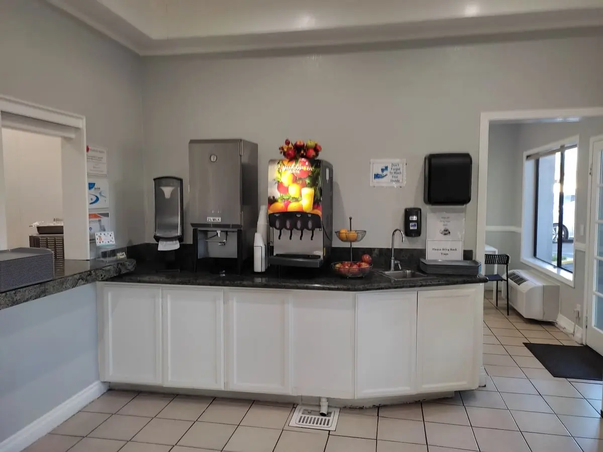 Counter with drinks (milk and juice) as well as fresh fruit for breakfast at Capri Suites Anaheim. On the left wall is a window at which guests can order hot breakfast items