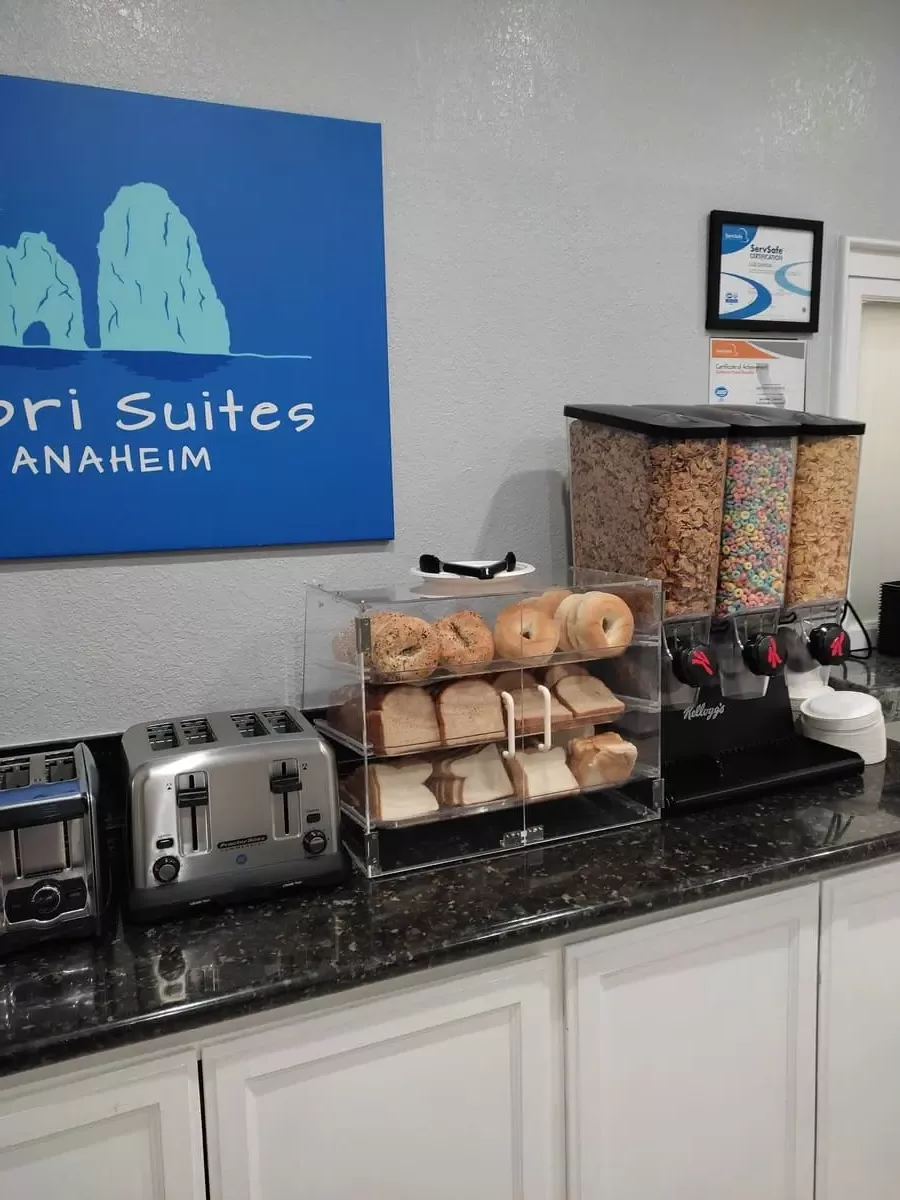 Part of the cold breakfast at Capri Suites Anaheim: Three containers with different creals, a small glass cabinet with different kinds of bagels and toast, and two toasters.