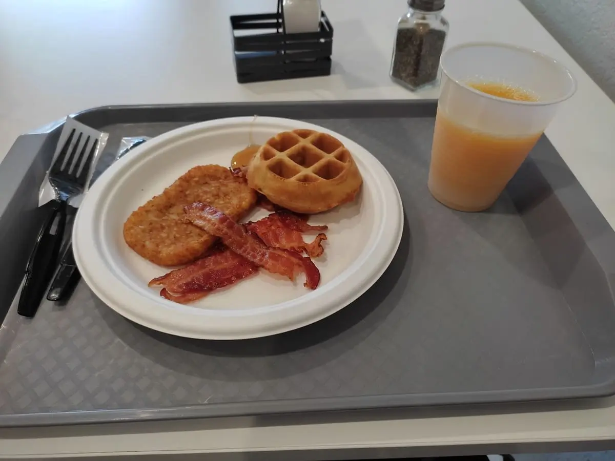 Tray with a cup with orange juice and paper plate with mini-waffle, bacon, and hash browns