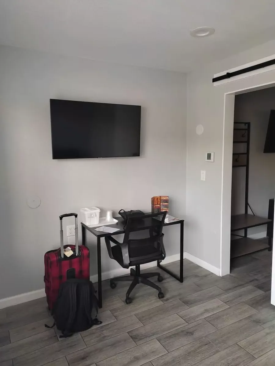Small desk and chair in a corner of the room. Next to it is a red and black suitcase and there is a TV above the desk.