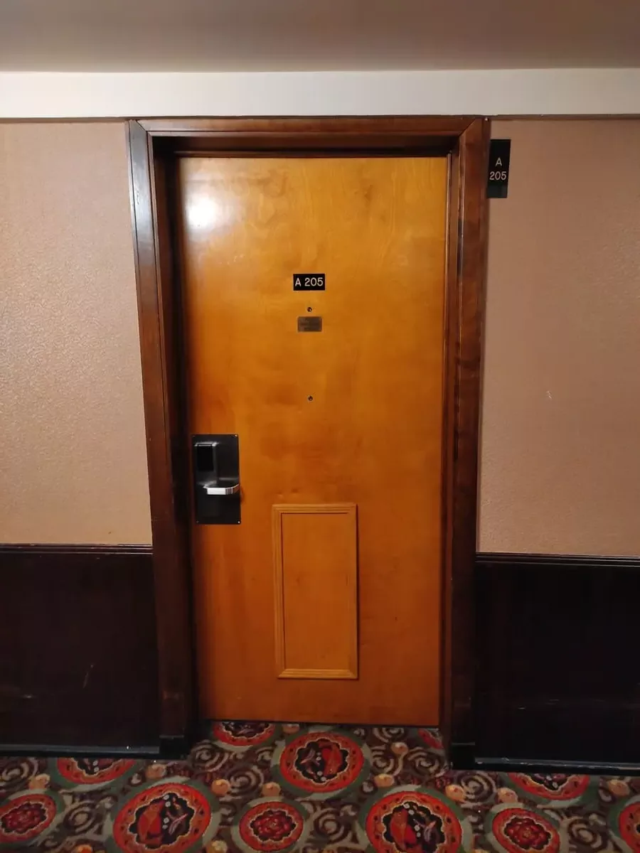 Wooden door to cabin A 205 at the end of the hallway where the wood paneling has ended and is replace by brown wallpaper.