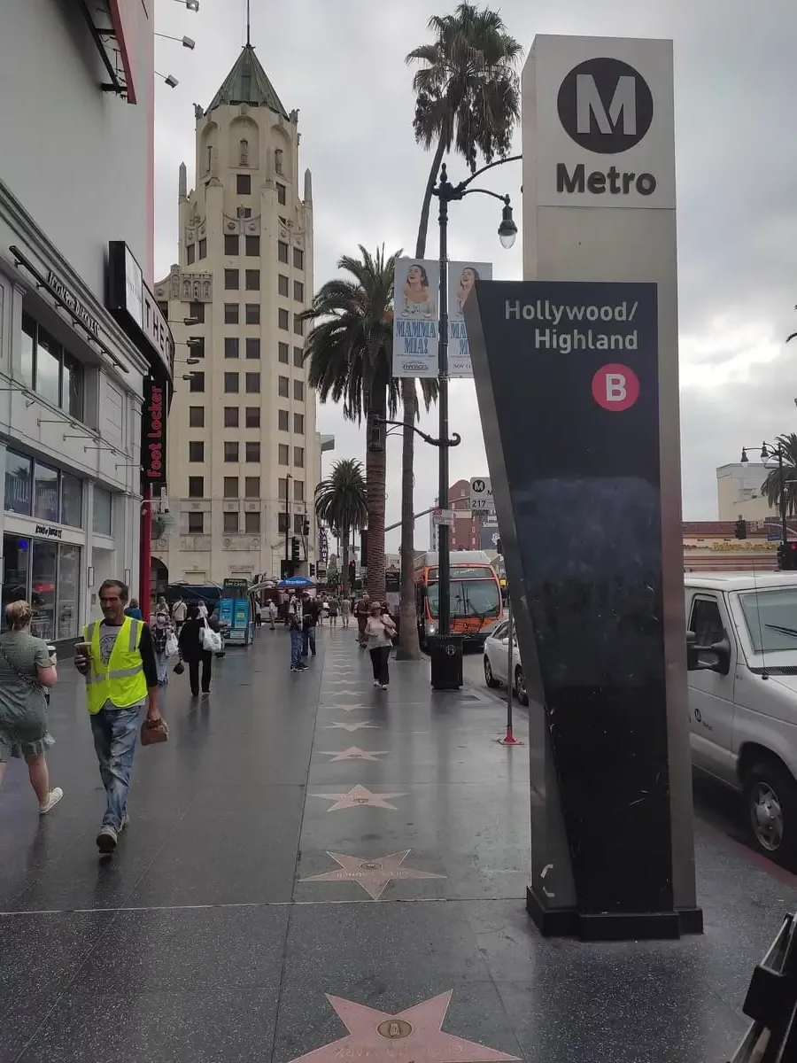 Busy Walk of Fame in Hollywood with pink stars on the ground. A large column clearly marks the entrance to the Hollywood/Highland stop of the Metro B line.