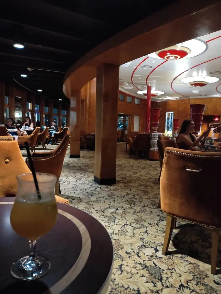 Queen Mary's Observation Lounge has a distinctive flowered carpet, large windows to the outside, small round tables, tan velvet chairs and impressive white and red Art deco lights