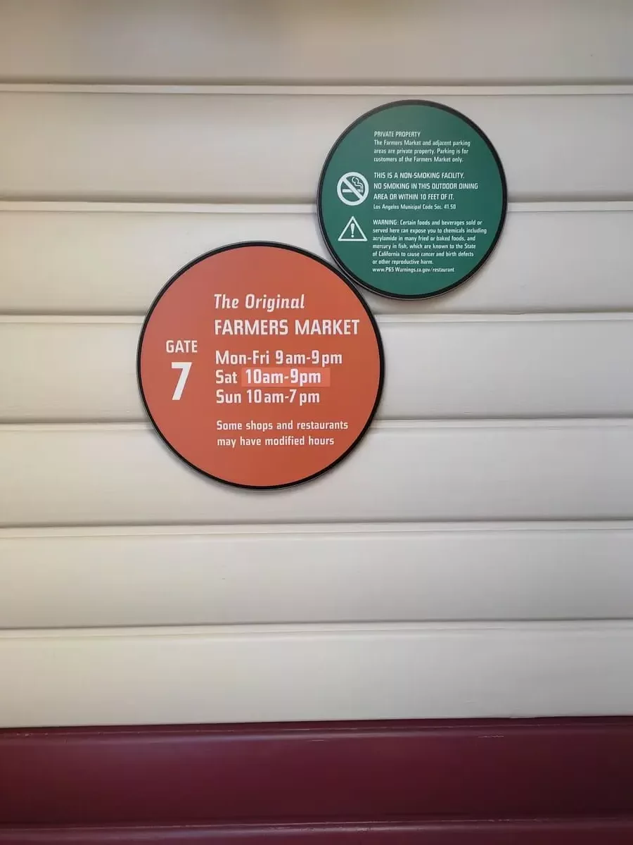 Clapboard wall with two round signs. One of which shows the opening times for The Original Farmers Market. The market closes surprisingly early.
