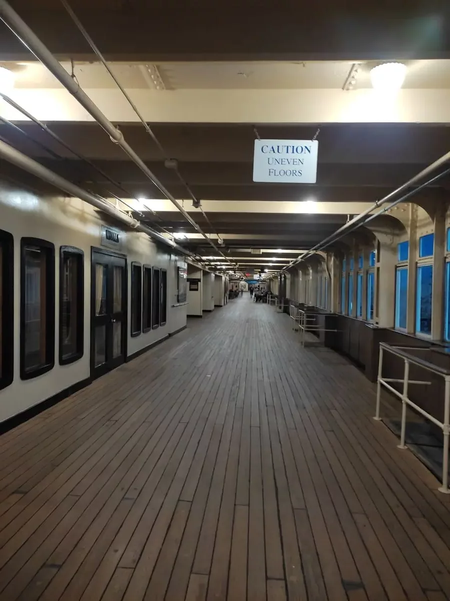 The empty promenade deck on Queen Mary has an eery vibe at night. It is enclosed with windows to the outside on the right and windows to various rooms on the left. The floor is made of wood.