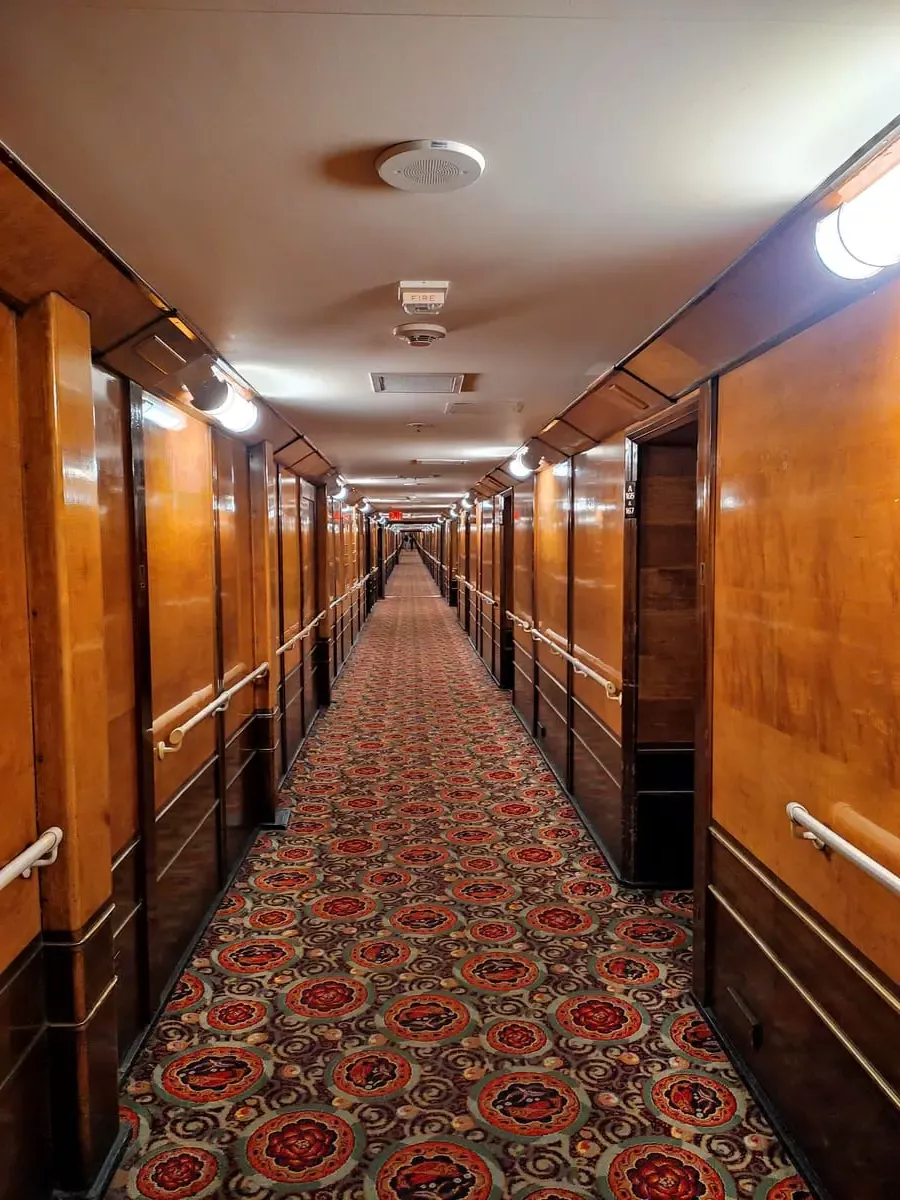 The long hallway on Queen Mary's A-deck has a distinctive red and green carpet with a flower pattern. The walls are paneled with a warm honey colored wood.