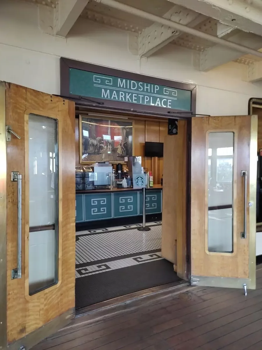 Two large wooden door open to Midship Marketplace on Queen Mary. The Starbucks property features a black and white tiled floor as well as a large painting over the counter