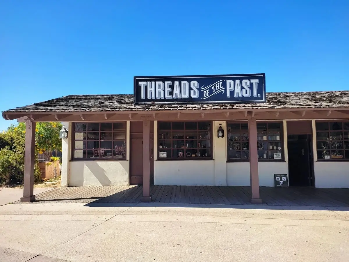 Low white building with old-fashioned window and a covered porch and the sign "Threads of the Past"