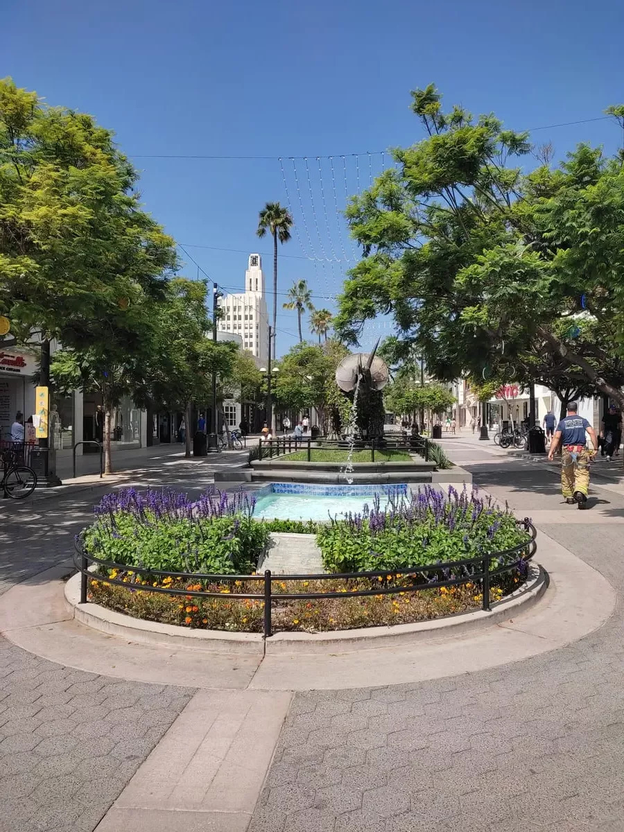 Santa Monica's Third Street Promenade is characterized by a wide pedestrian street with colorful flowers, fountains, and large dino sculptures.