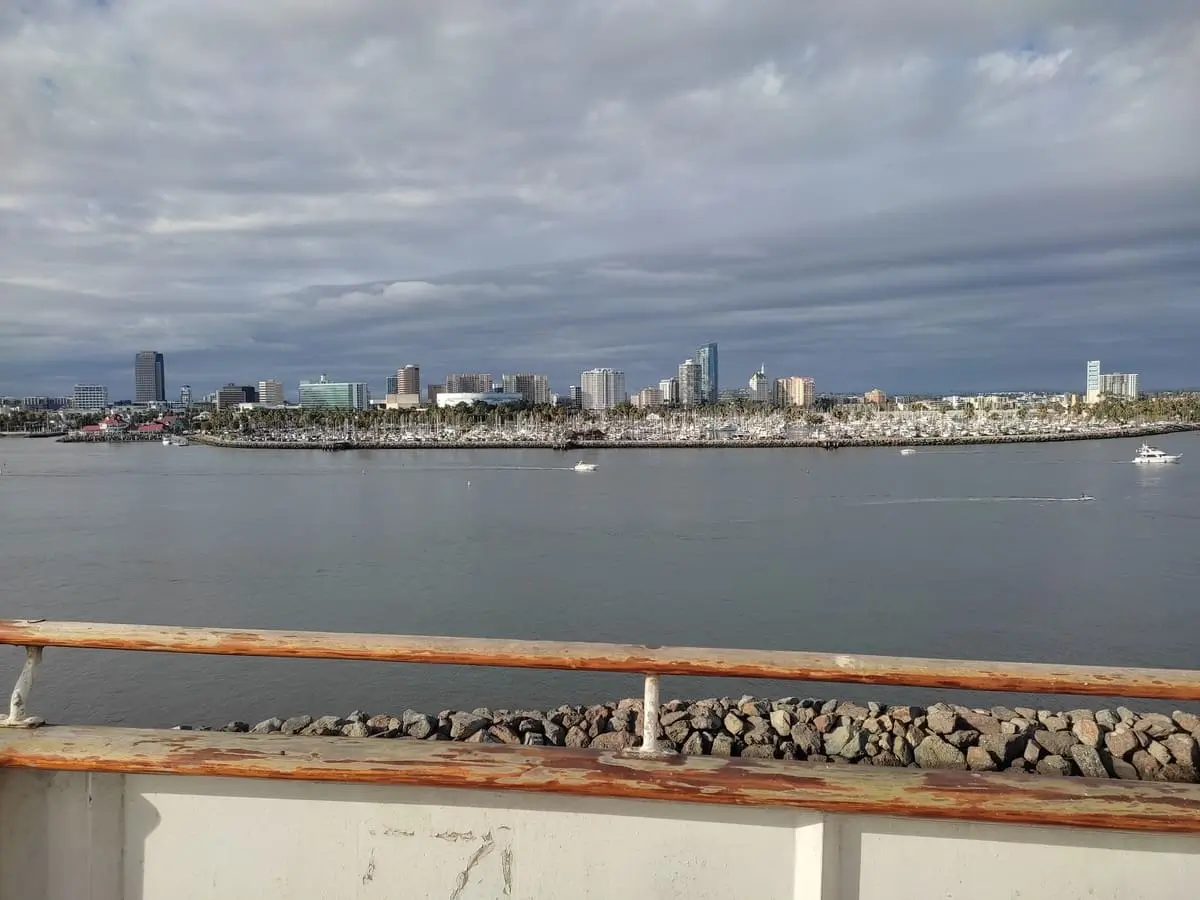 Over the chipped wooden railing of hotel ship Queen Mary you can see the Long Beach skyline across the bay. Small yachts are moored in front of the line of skyscrapers and several boats are out on the water.