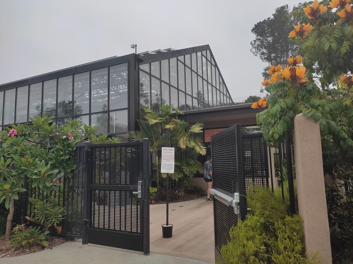 The path through the large metal gate leads to the ticket counter for the San Diego Botanic Garden in Enicinitas. Next to it is a large greenhouse.