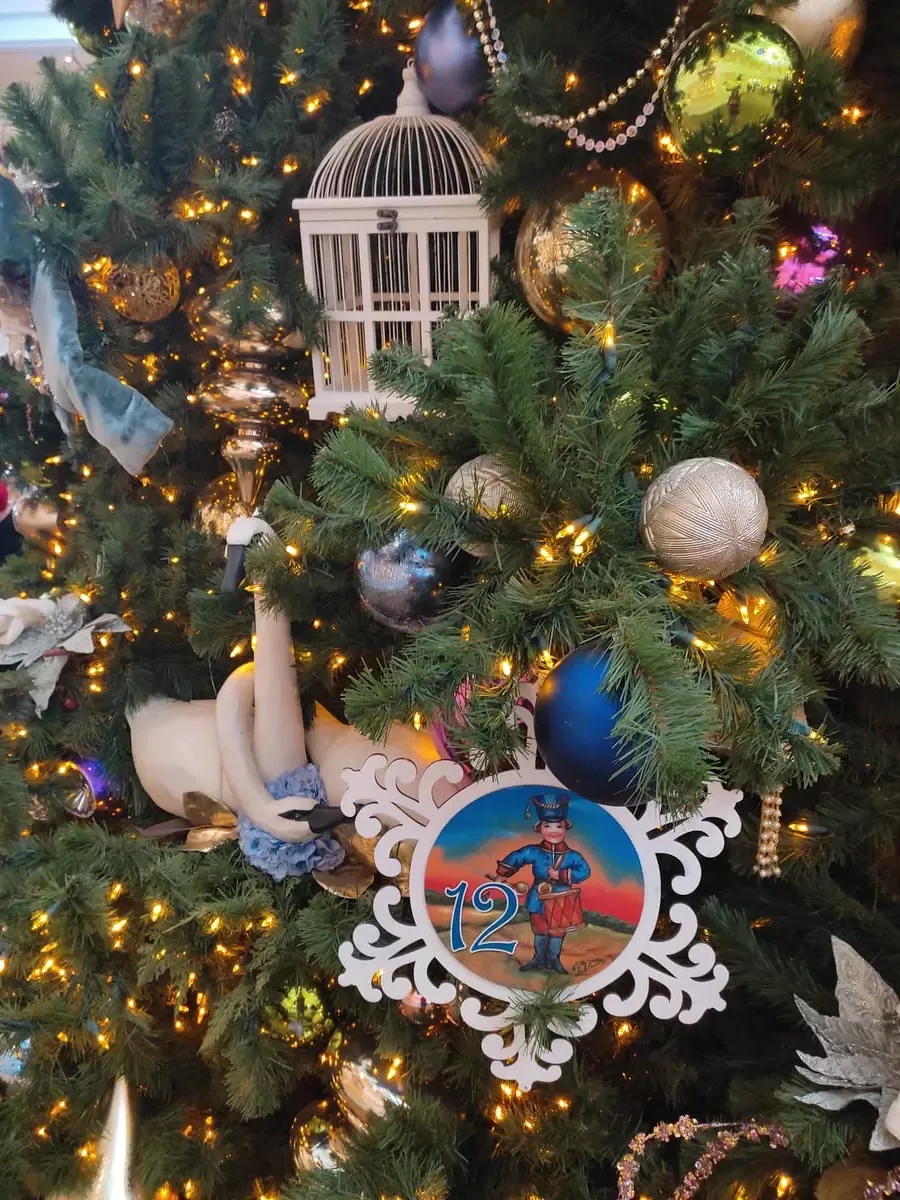 Close-up of Christmas tree decorations with colors mainly white and blue. There are two swans, a birdcage, and a picture of a drummer with the number 12.