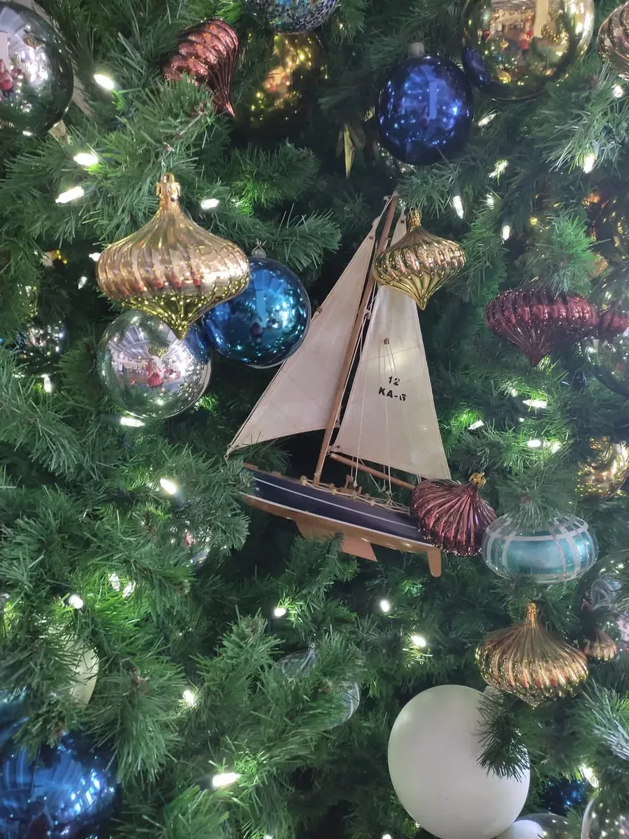 Close-up of Christmas tree decorations with colors mainly blue, burgundy, and gold. There are metallic ornaments and a model of a sail boat.